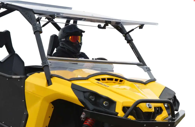 A rider in black motocross gear and red-tinted goggles sits inside a yellow vehicle equipped with a Can-Am Commander Scratch Resistant Flip Windshield (2011-2020), featuring a roll cage, against a plain white background.