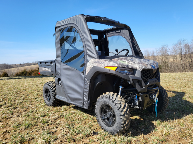 A CFMoto UForce 600 with a Doors-Rear Window Combo is parked on grassy, uneven ground under a clear blue sky. Bare trees and rolling hills are in the background. The utility vehicle features rugged tires and a front winch.