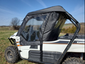 A Kawasaki Teryx 800 (2-Seater) with a Modular Full Cab for Hard Windshield, featuring black trim and clear windows, is parked on grassy terrain with open fields, trees, and a blue sky in the background.