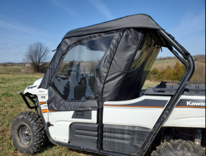 A Kawasaki Teryx 800 (2-Seater) with a Modular Full Cab for Hard Windshield, featuring black trim and clear windows, is parked on grassy terrain with open fields, trees, and a blue sky in the background.