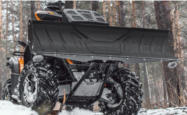 A low-angle close-up of an orange ATV with rugged tires and a Rival Powersports Universal ATV Snow Plow Kit (stamped steel blade, 48/54/60 with mount) is parked in a snowy pine forest, its wheels and plow covered in snow and mud.