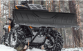 A low-angle close-up of an orange ATV with rugged tires and a Rival Powersports Universal ATV Snow Plow Kit (stamped steel blade, 48/54/60 with mount) is parked in a snowy pine forest, its wheels and plow covered in snow and mud.