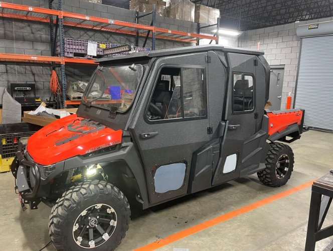 A GC1K Crew Armor Tech Enclosure, a rugged black and red off-road utility vehicle with large tires and an enclosed cabin, is parked in an industrial warehouse with metal shelves, boxes, and tools visible in the background.