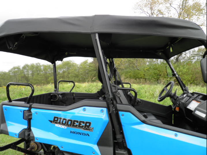 A Honda Pioneer 1000-5 Soft Top side-by-side utility vehicle with a black roof and open sides is parked on grass, with seats and steering wheel visible, set against a backdrop of trees and cloudy sky.