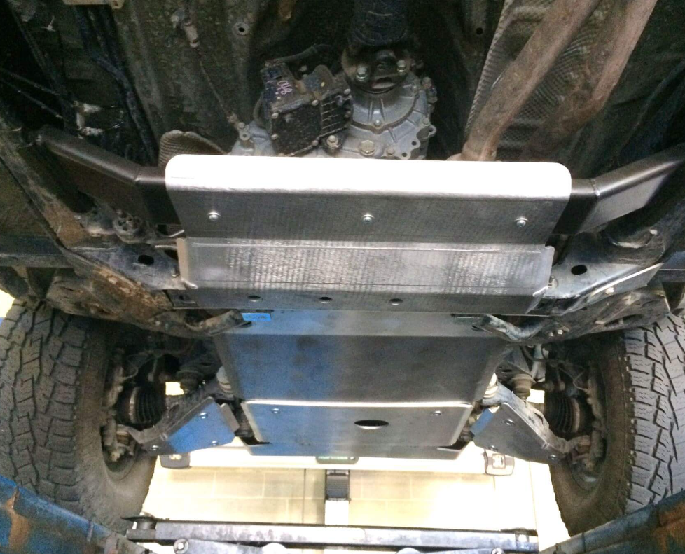 Underneath a raised vehicle, the 9-Piece Complete Aluminum Skid Armor Kit for Toyota Tacoma is shown mounted, protecting the engine and transmission. Tires and suspension are visible at the sides, with some dirt on nearby components.