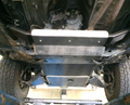 Underneath a raised vehicle, the 9-Piece Complete Aluminum Skid Armor Kit for Toyota Tacoma is shown mounted, protecting the engine and transmission. Tires and suspension are visible at the sides, with some dirt on nearby components.
