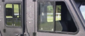 Close-up of a matte black armored vehicle featuring Sliding Glass Window Replacement Armor Tech with square windows. The doors have visible rivets and reflect green grass and trees. The image is slightly blurry and only part of the vehicle is visible.
