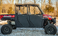 A red and black off-road vehicle with large tires, robust suspension, and Polaris Ranger 1000 Aluminum Lower Doors (2020+) by SuperATV is parked on snow-dusted ground among leafless trees in a wooded area on a clear day.