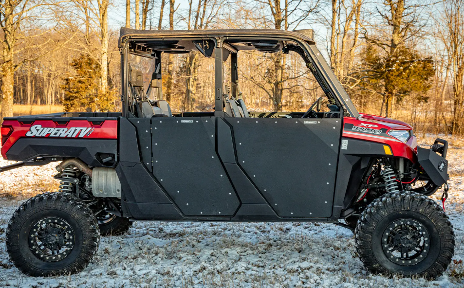 A red and black off-road vehicle with large tires, robust suspension, and Polaris Ranger 1000 Aluminum Lower Doors (2020+) by SuperATV is parked on snow-dusted ground among leafless trees in a wooded area on a clear day.