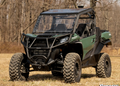 A green and black off-road vehicle with a Can-Am Maverick Trail Scratch Resistant Flip Windshield, large rugged tires, roll cage, and two seats is parked on dry grass, with leafless trees and a clear sky in the background.