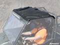A person in an orange shirt and black helmet with goggles drives a utility vehicle equipped with a Polaris Ranger XP Plastic Roof and metal roll cage through dry grassy terrain. The SuperATV logo is displayed in the bottom right corner.