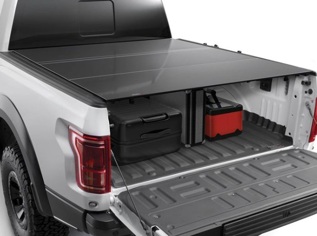 A 2014-18 Chevy Silverado 6ft 6in box is shown with the Alloycover Hard Folding Bed Cover (black) partially open, revealing two black suitcases and a red and black toolbox in the bed; rear tires and taillight are visible.