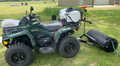 A green Can-Am Outlander ATV is parked on grass with a white plastic tank sprayer on the rear rack. Attached to the back is a PackerMaxx ATV/UTV Cultipacker Heavy Duty 4 Ft Pro Series, likely for lawn care tasks. A building appears in the background.