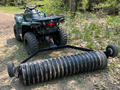 A green ATV is parked on a dirt path in the woods, hitched to a PackerMaxx Heavy Duty 6 Ft Pro Series ATV/UTV Cultipacker, with two small wheels and black corrugated finish, surrounded by trees and foliage in daylight.