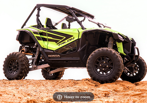 A Honda Talon with dark tinted lower door inserts is parked on sandy terrain. The bright green and black off-road vehicle features large rugged tires, two seats in an open cockpit, and a protective roll cage, viewed from a low angle against a white backgr