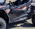 Close-up of black and gray Polaris RZR 900/1000/Turbo featuring lower door inserts with red and white accents, rugged tires, visible seats, and prominent Polaris logo. The off-road vehicle is parked outdoors.