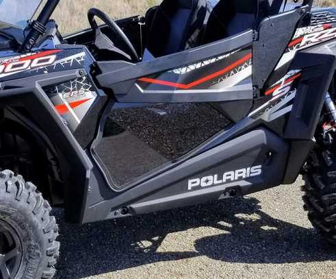 Close-up of black and gray Polaris RZR 900/1000/Turbo featuring lower door inserts with red and white accents, rugged tires, visible seats, and prominent Polaris logo. The off-road vehicle is parked outdoors.