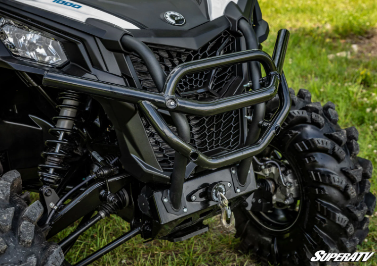 Close-up of a rugged black Can-Am Maverick Trail Winch Mounting Plate with +3500lb winch on an ATV with large knobby tires. The ATV is parked on grass, and SuperATV branding appears in the lower right corner.