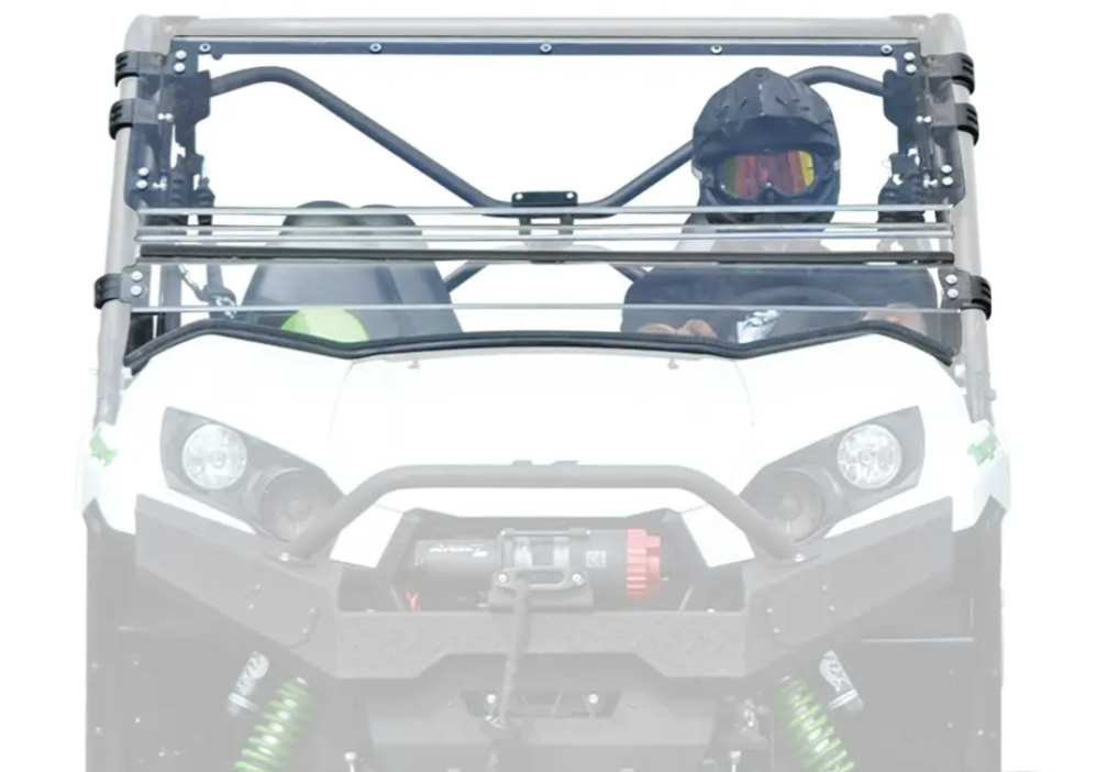 A person in a black helmet and goggles sits behind a Kawasaki Teryx 750 / 800 Scratch Resistant Flip Windshield in the driver’s seat of a white UTV with roll cage, winch, and visible suspension, shown from the front.