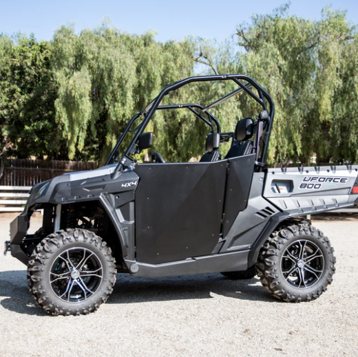 A CF Moto UFORCE 800 with Half Doors by Dirt Specialties, black rims, rugged off-road tires, and roll cage is parked on gravel. The side-by-side seats two, with green trees in the background on a sunny day.