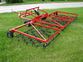 The Universal ATV/UTV Harrow - Heavy Duty Commercial Grade, a large red metal drag harrow with chain tines, black wheels, and a hitch for vehicle attachment, sits on green grass near a paved road and trees.