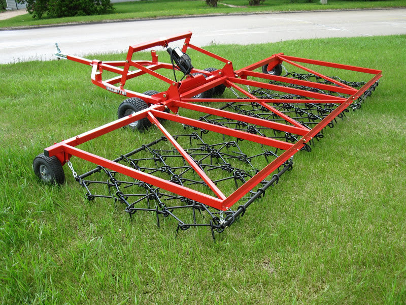 The Universal ATV/UTV Harrow - Heavy Duty Commercial Grade, a large red metal drag harrow with chain tines, black wheels, and a hitch for vehicle attachment, sits on green grass near a paved road and trees.