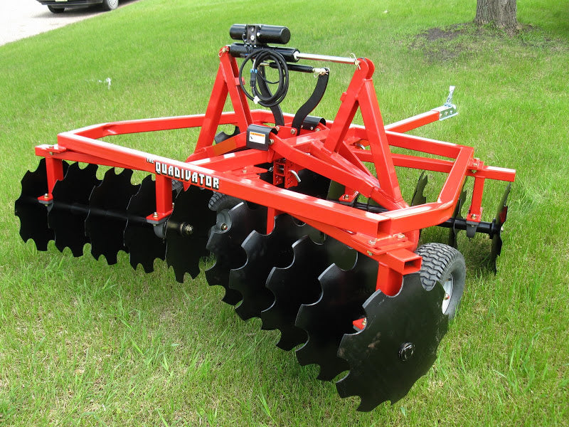 A Universal Atv/Utv Tandem Disc - Heavy Duty Commercial Grade sits on green grass, featuring multiple black scalloped discs in rows, a tractor hitch, hydraulic hoses, and wheels. A tree and part of a parked vehicle are in the background.