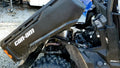 A Can-Am Defender equipped with the Bed Lift – KYB Lift Cylinder Actuator/Electric Lift Kit (Linear Actuator) MMP4-B1B250BB-DHR shows its rear cargo bed elevated, revealing engine parts and a large tire on an outdoor gravel surface.