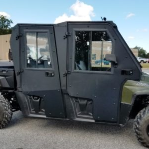 A POLARIS RANGER XP800 CREW PROTECTOR CAB, featuring two reinforced doors, armored panels, and large off-road tires, is parked on pavement near an industrial building with trees and a partly cloudy sky in the background.