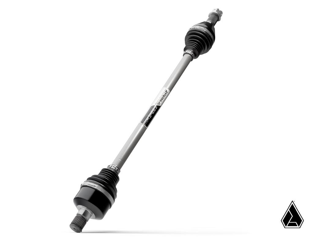 The Assault Industries MAS-223 Phoenix Axle for Can-Am Maverick R is shown on a white background, with black rubber boots on each end of its metallic rod, a barcode label, and a small black geometric logo in the lower right corner.