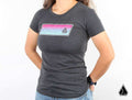 A person stands against a white background wearing the Assault Industries Miami Vice Womens Tee, a fitted dark gray shirt with a colorful horizontal stripe and small geometric logo, paired with blue jeans and a black smart watch.