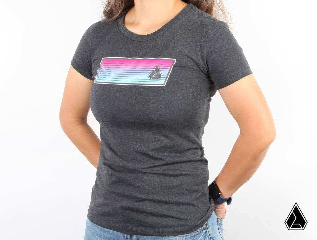 A person stands against a white background wearing the Assault Industries Miami Vice Womens Tee, a fitted dark gray shirt with a colorful horizontal stripe and small geometric logo, paired with blue jeans and a black smart watch.
