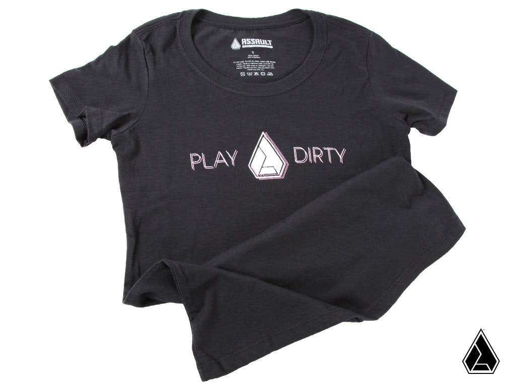 The Assault Industries 2021 Play Dirty Womens Tee is a black short-sleeve shirt featuring “PLAY DIRTY” in white and pink across the chest, separated by a geometric logo. The tee is shown laid flat on a white background with slight folds at one sleeve and 