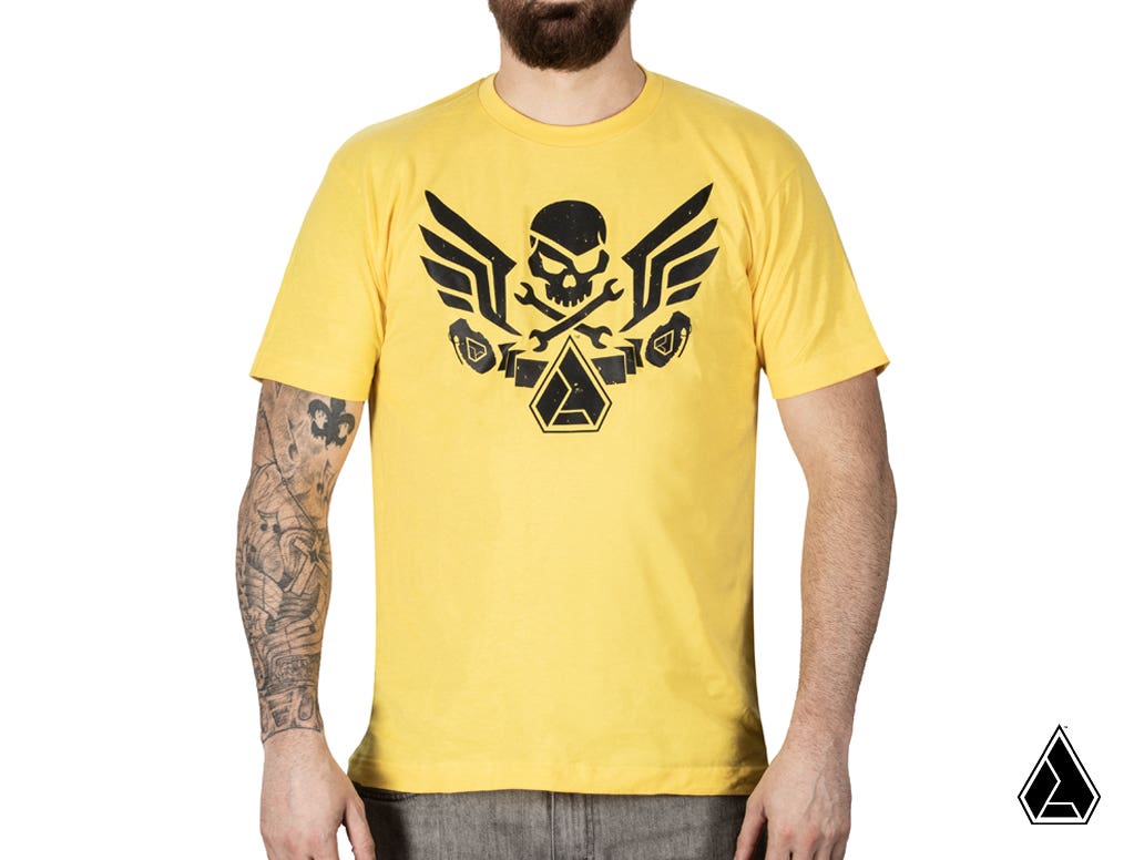 A person with a beard and a tattoo sleeve wears the Assault Industries Raider Yellow T-Shirt, featuring a black skull, wings, and crossed wrenches graphic on the chest. A small black logo appears at the bottom right corner of the image.