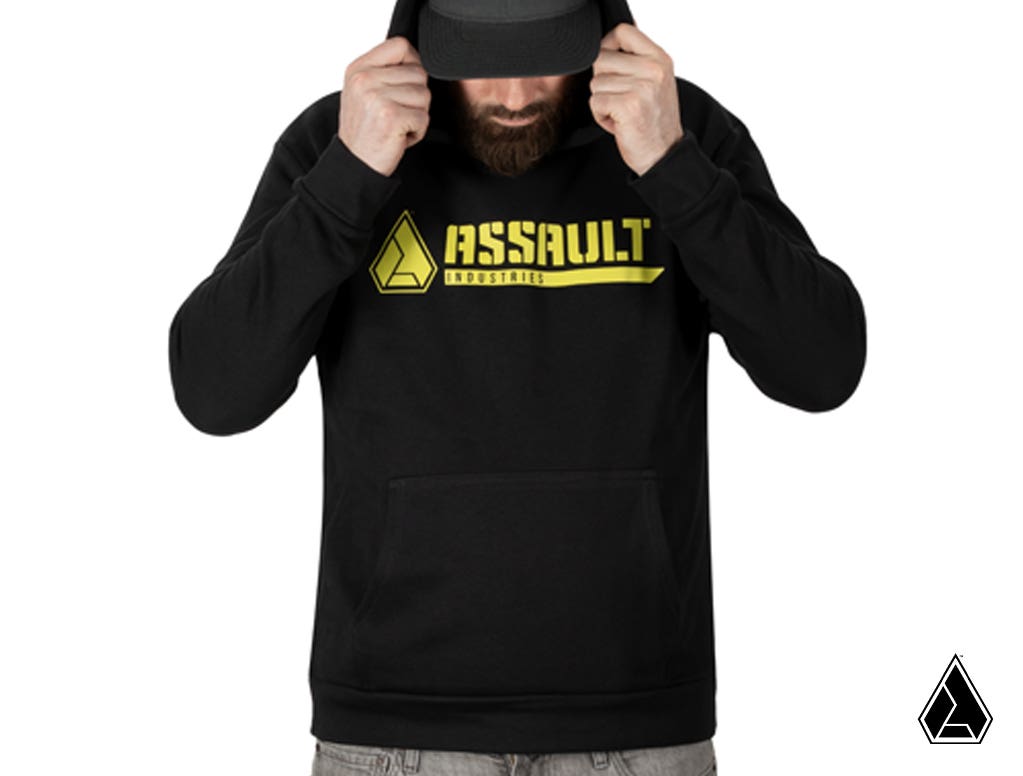 A bearded man in a black Assault Industries Bomb Hoodie with a yellow logo across the chest and cap looks down, holding the hood with both hands. The background is white, with a small triangular logo in the bottom right corner.