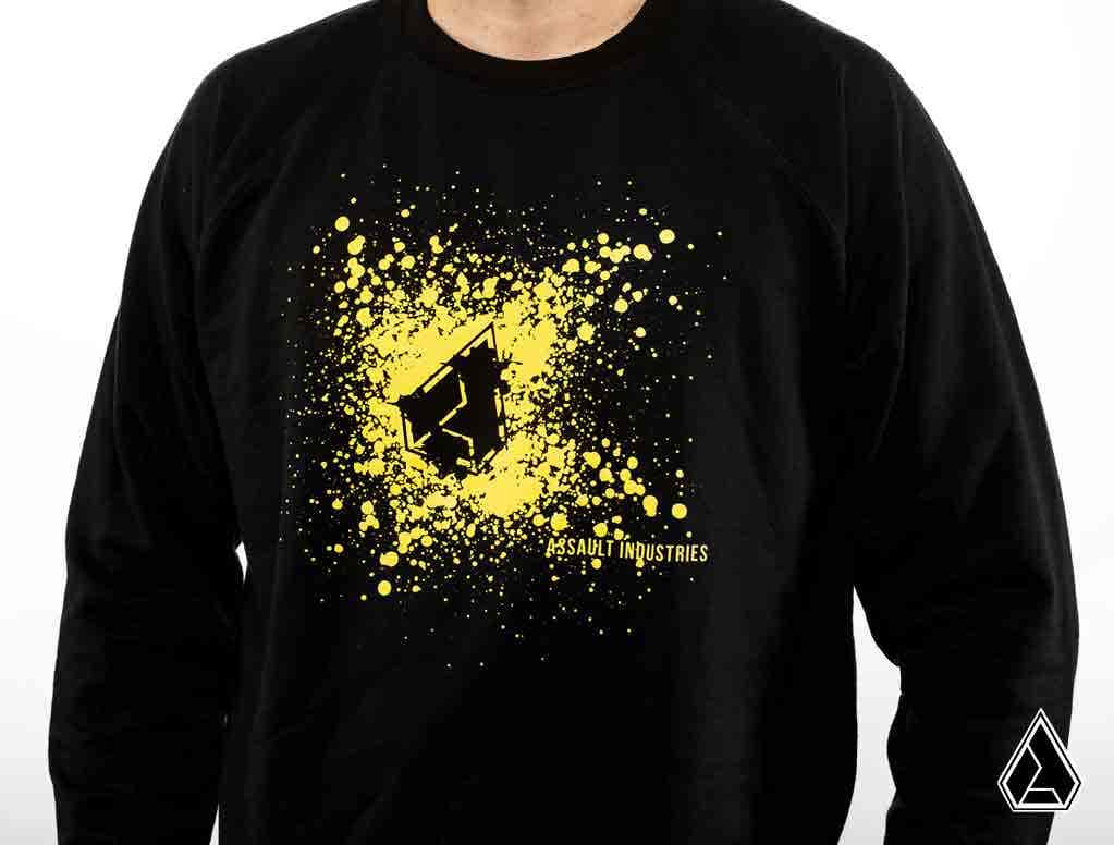 Someone wears the Assault Industries Impact Crewneck Sweatshirt, featuring a black base with a yellow splatter design and a stylized wolf head logo centered above small ASSAULT INDUSTRIES text. The wearer’s face is not visible.