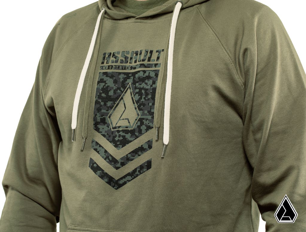 A person wears the Assault Industries Chevron Hoodie in olive green with white drawstrings, a geometric digital camo logo and ASSAULT on the chest, raglan sleeves, and a small white triangle logo at the bottom right.