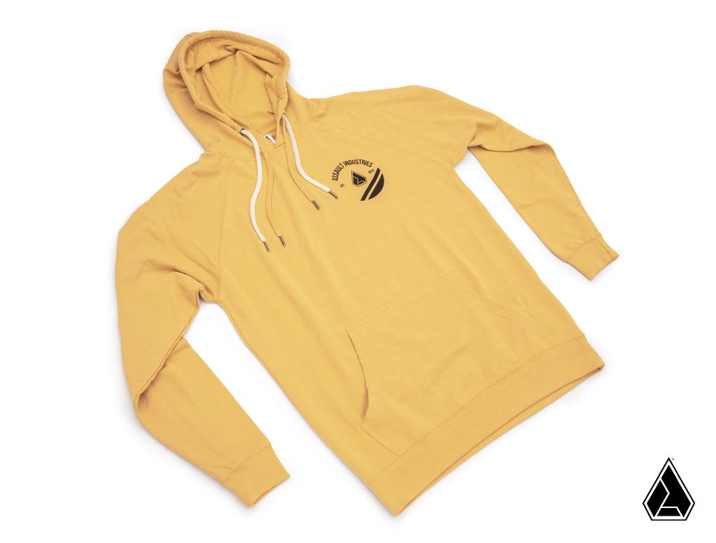 The Assault Industries Gold Hoodie is a mustard yellow hooded sweatshirt with white drawstrings, a front pocket, and a small black circular mountain logo on the left chest. It’s shown flat on a white background with a black triangle logo at the bottom rig