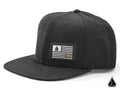 The Assault Industries Snapback Hat is black with a flat brim and features a front patch of a stylized flag with stripes and a small geometric logo. It’s displayed on a plain white background, plus the matching geometric logo in the bottom right corner.
