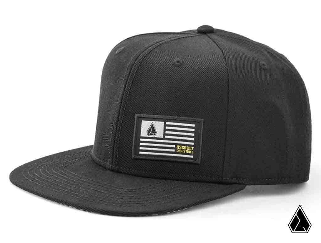 The Assault Industries Snapback Hat is black with a flat brim and features a front patch of a stylized flag with stripes and a small geometric logo. It’s displayed on a plain white background, plus the matching geometric logo in the bottom right corner.