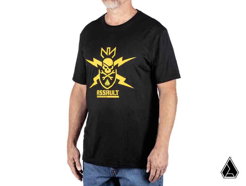 A bearded man stands against a white background, wearing the Assault Warning Label Mens T-Shirt featuring a bold yellow skull graphic with tools, lightning bolts, and crown. ASSAULT appears below the design along with a small geometric logo in the corner.