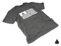 A heather gray Assault Industries Flag Mens T-Shirt lies flat, featuring a white wavy stripe graphic and pentagonal logo at the top left with RESOLUTE below, plus a matching small logo at the lower right corner.
