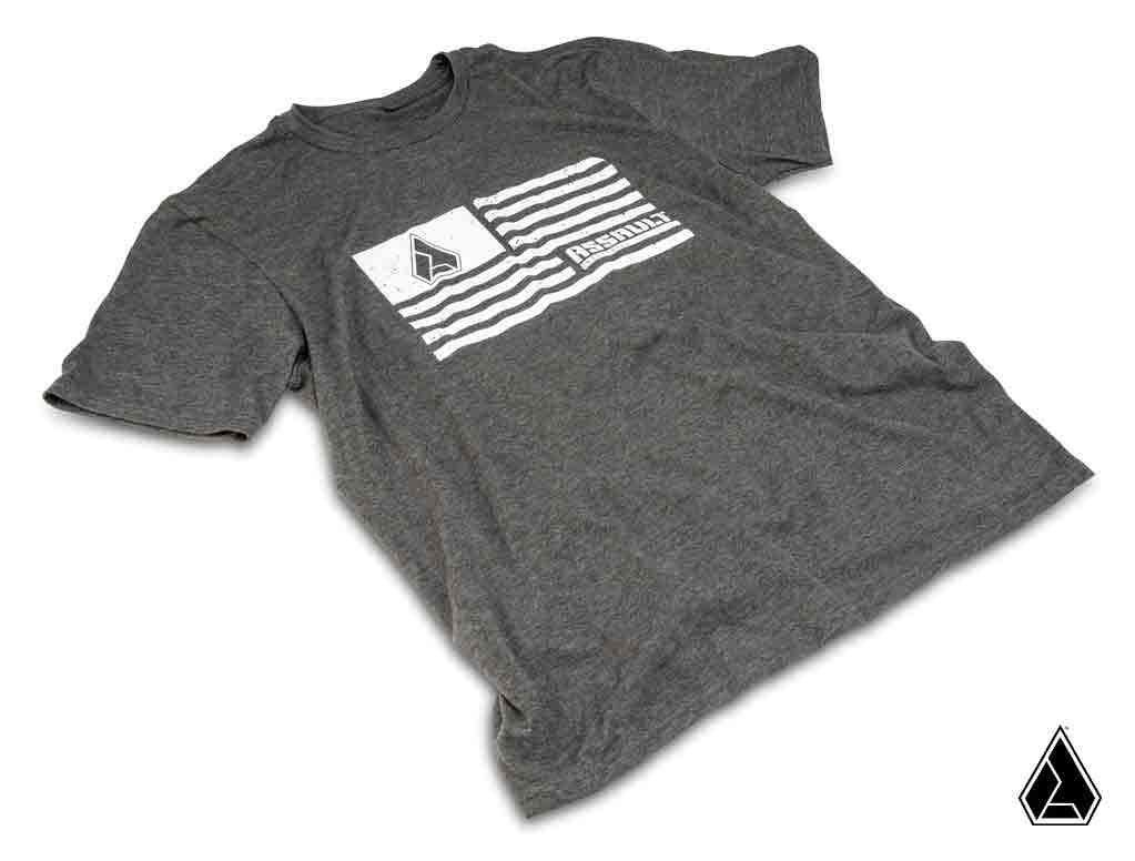 A heather gray Assault Industries Flag Mens T-Shirt lies flat, featuring a white wavy stripe graphic and pentagonal logo at the top left with RESOLUTE below, plus a matching small logo at the lower right corner.