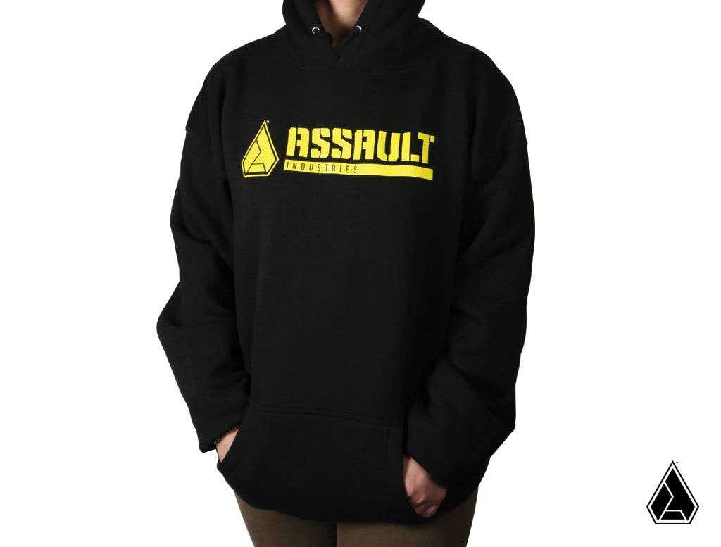 A person wears the Assault Industries Classic Logo Hoodie, featuring bold yellow “ASSAULT Industries” text and logo on the chest. Their hands are in the front pocket. The image has a plain white background with a small triangular logo at the bottom right.