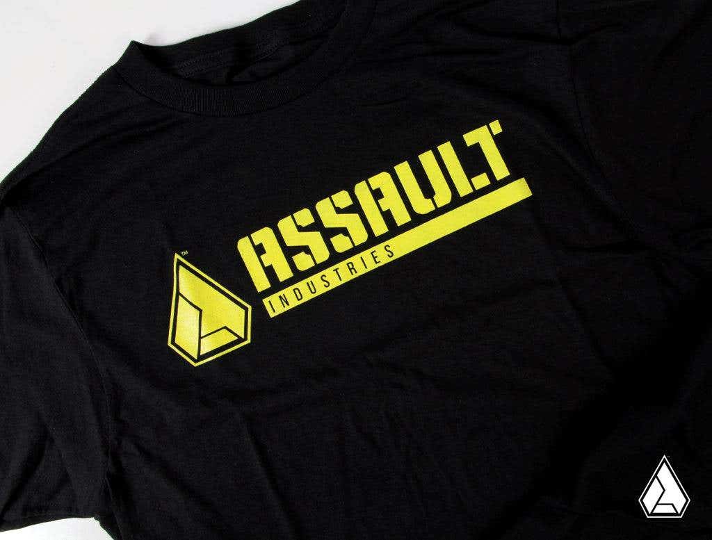 The Assault Industries Classic Logo Tee is a black t-shirt featuring “ASSAULT” in bold yellow letters above “INDUSTRIES,” with a yellow geometric triangle logo on the left and a smaller white version in the lower right corner.