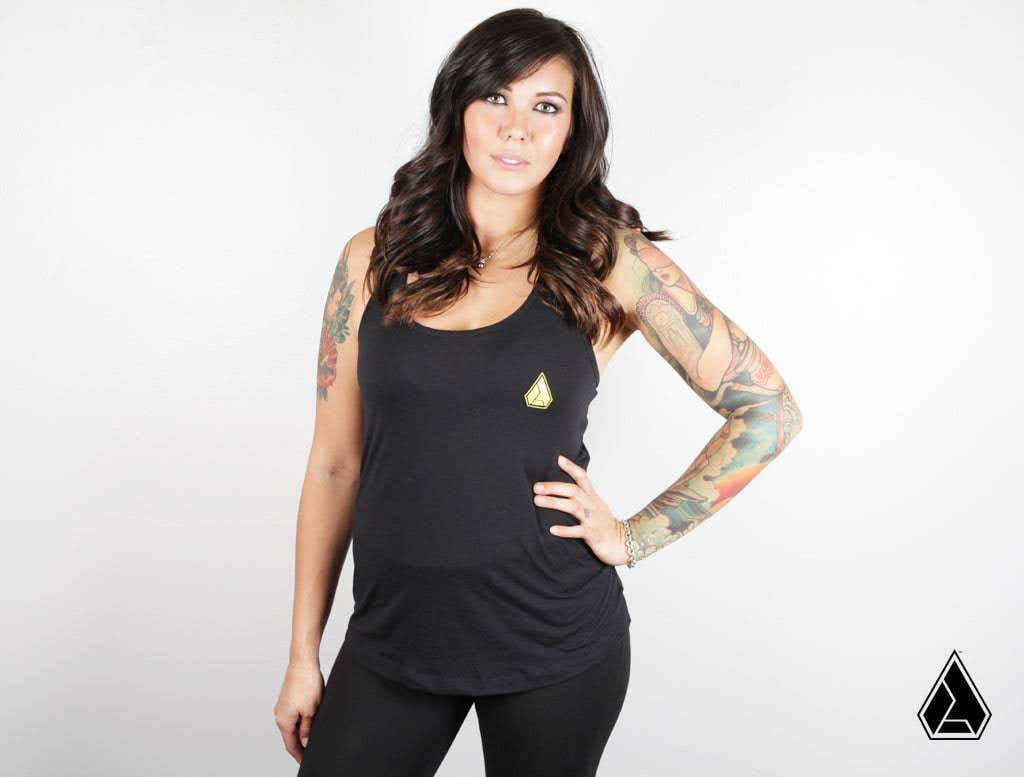 A woman with long dark wavy hair and arm tattoos stands against a white background, confidently wearing the Assault Industries Womens Iconic Logo Tank Top and black leggings. Her left hand is on her hip as she faces the camera.