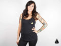 A woman with long dark wavy hair and arm tattoos stands against a white background, confidently wearing the Assault Industries Womens Iconic Logo Tank Top and black leggings. Her left hand is on her hip as she faces the camera.
