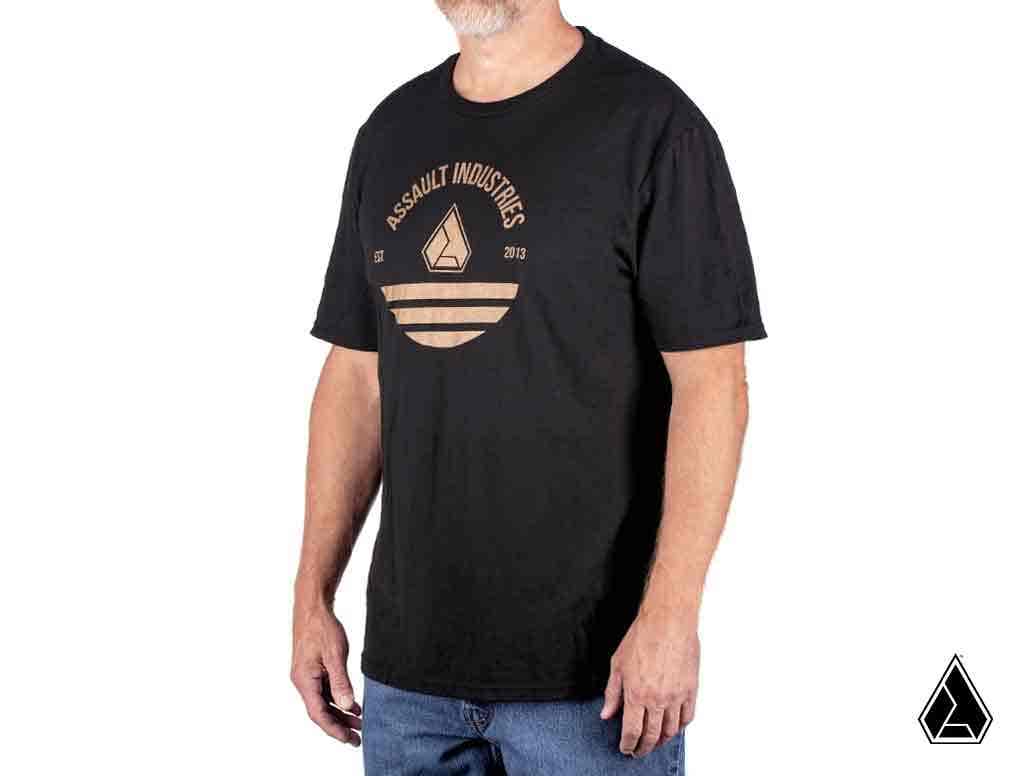 A man with a short gray beard wears the Assault Golden Sunset Mens T-Shirt, featuring a round graphic logo reading Assault Industries Est. 2013 above a geometric shape and stripes. He stands slightly turned left against a plain white background. A small l