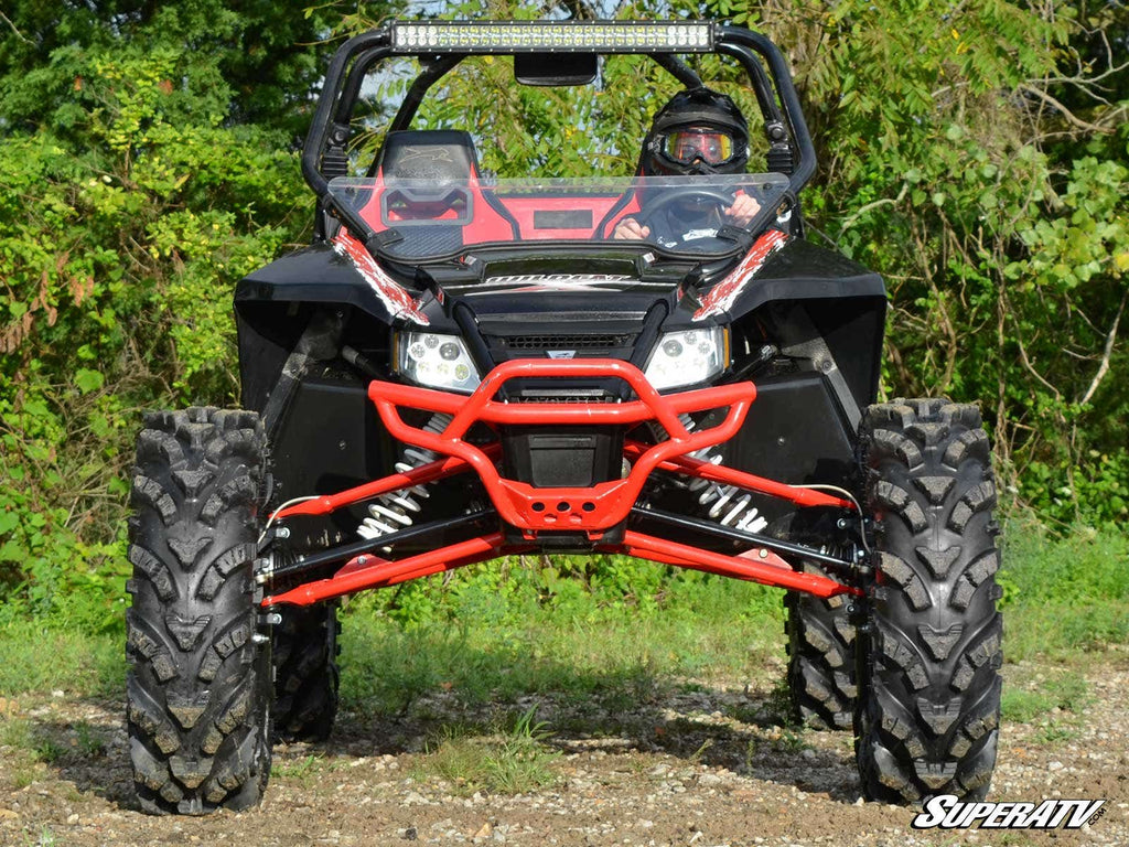 A rugged ATV equipped with the Arctic Cat Wildcat 4 Portal Gear Lift faces forward on rocky ground. Its large, knobby tires and raised suspension stand out as a helmeted driver sits inside, with green trees in the background.
