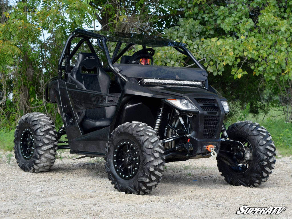 An Arctic Cat Wildcat Trail equipped with a 5 Long Travel Kit is parked on gravel. Its black body features angular panels, rugged tires, and a protective roll cage, with dense green foliage in the background. SuperATV logo appears lower right.